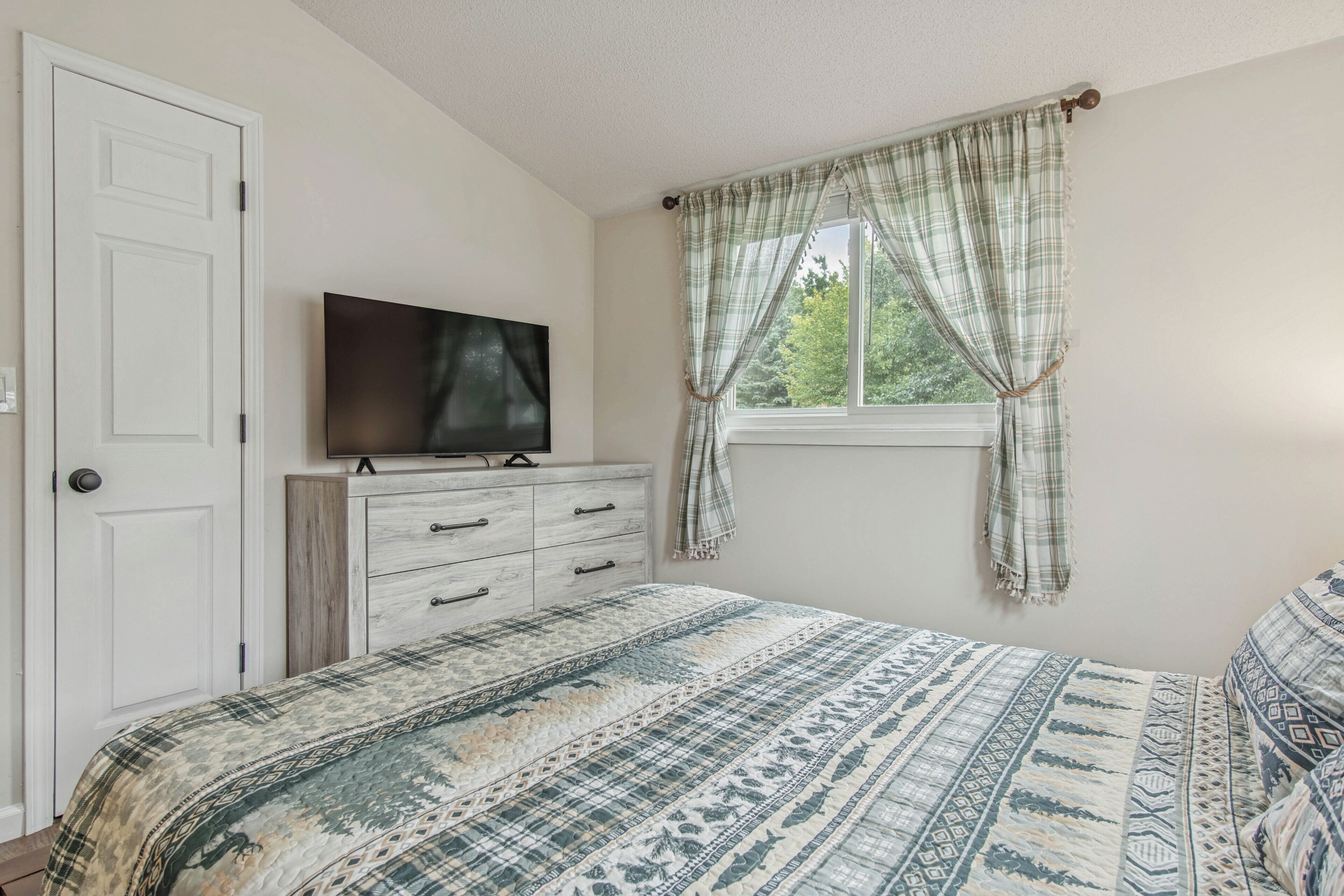Een slaapkamer met een bed, een kast, een televisietoestel bovenop de kast, en een raam met gordijnen.