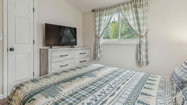 A bedroom with a bed, a dresser, a television on top of the dresser, and a window with curtains. - Lake It Easy - NNY Waterfront Home on Lake Ontario (Dexter)