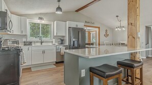 A modern kitchen with a central island, bar stools, and a view of the dining area. - Lake It Easy - NNY Waterfront Home on Lake Ontario (Dexter)