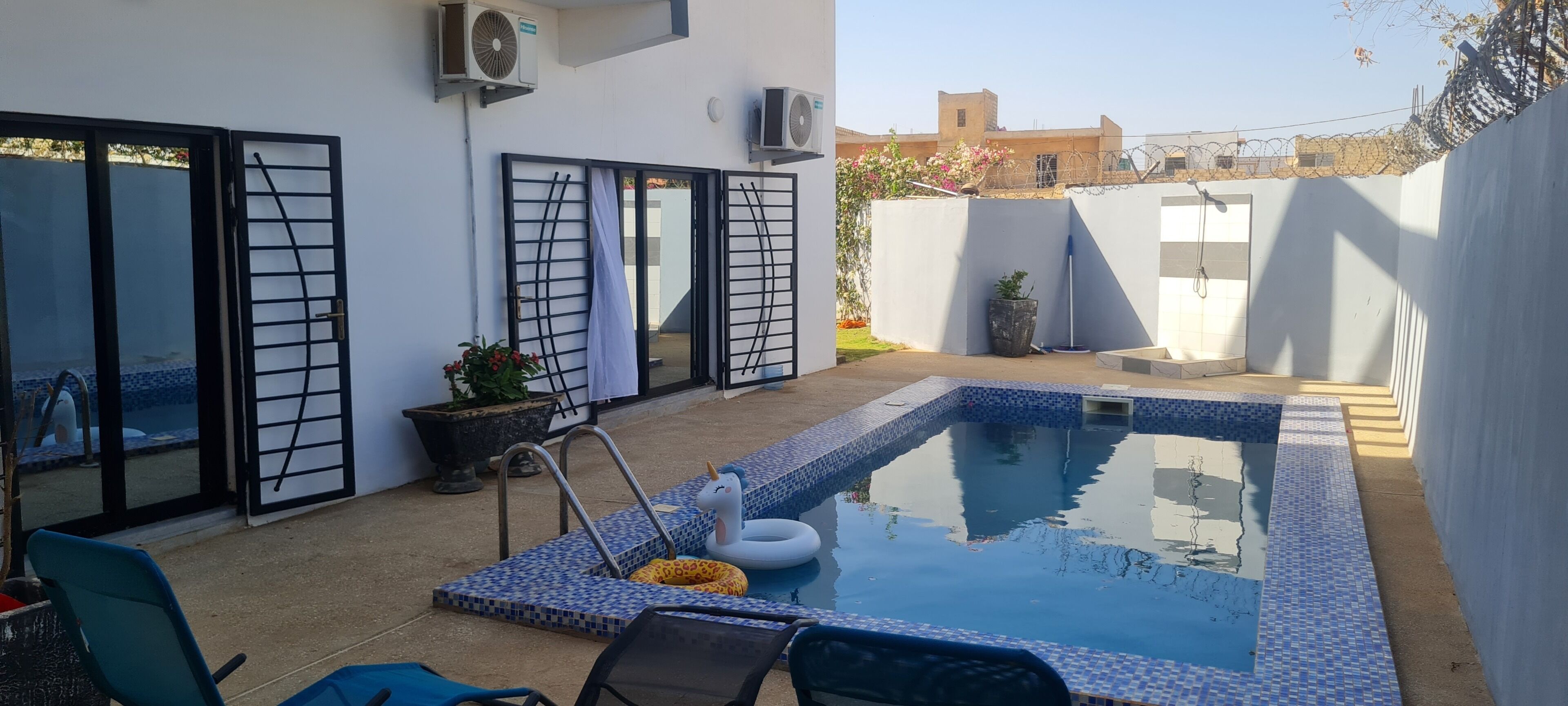 A swimming pool with a tiled edge, a lounge chair, a potted plant, and a barbed wire fence.