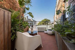 Ein schmaler Balkon mit Gartenmöbeln, darunter ein weißer Stuhl und ein Tischchen, umgeben von Blumentöpfen und einem Holenzaun.
