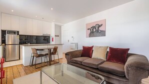 Una sala de estar moderna con una mesa de centro de vidrio, un sofá marrón con cojines, y una zona de cocina con armarios blancos y una encimera negra.
