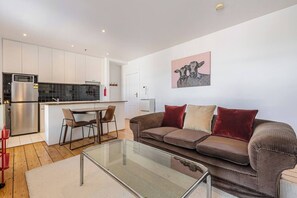 Ein modernes Wohnzimmer mit einem Glastisch, einem braunen Sofa mit Kissen und einer Küchenzeile mit weißen Schränken und einer schwarzen Arbeitsplatte.