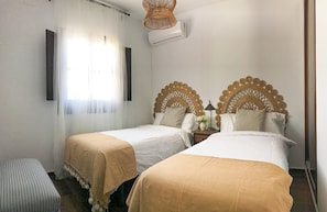 A hotel room with two single beds, a bedside table with a lamp, a window with sheer curtains, and an air conditioning unit on the wall.
