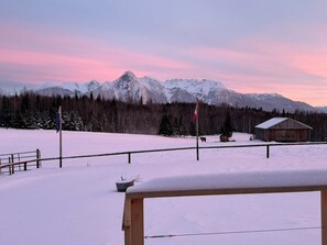 A snowy landscape with a wooden cabin and a flagpole in the foreground, and snow-covered mountains in the background.