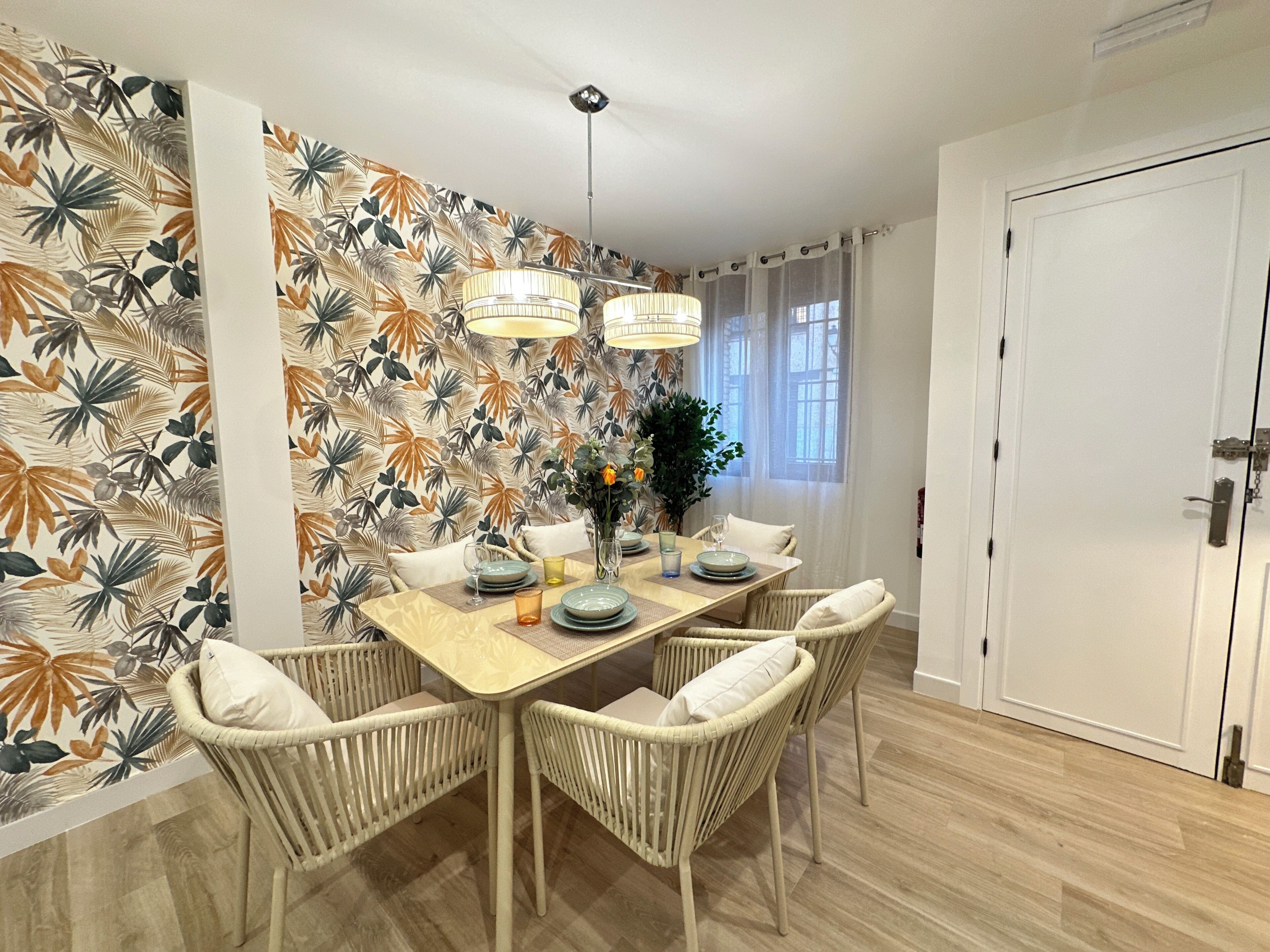 A dining area with a table set for four, surrounded by wicker chairs, a patterned wall, and a door to the right.