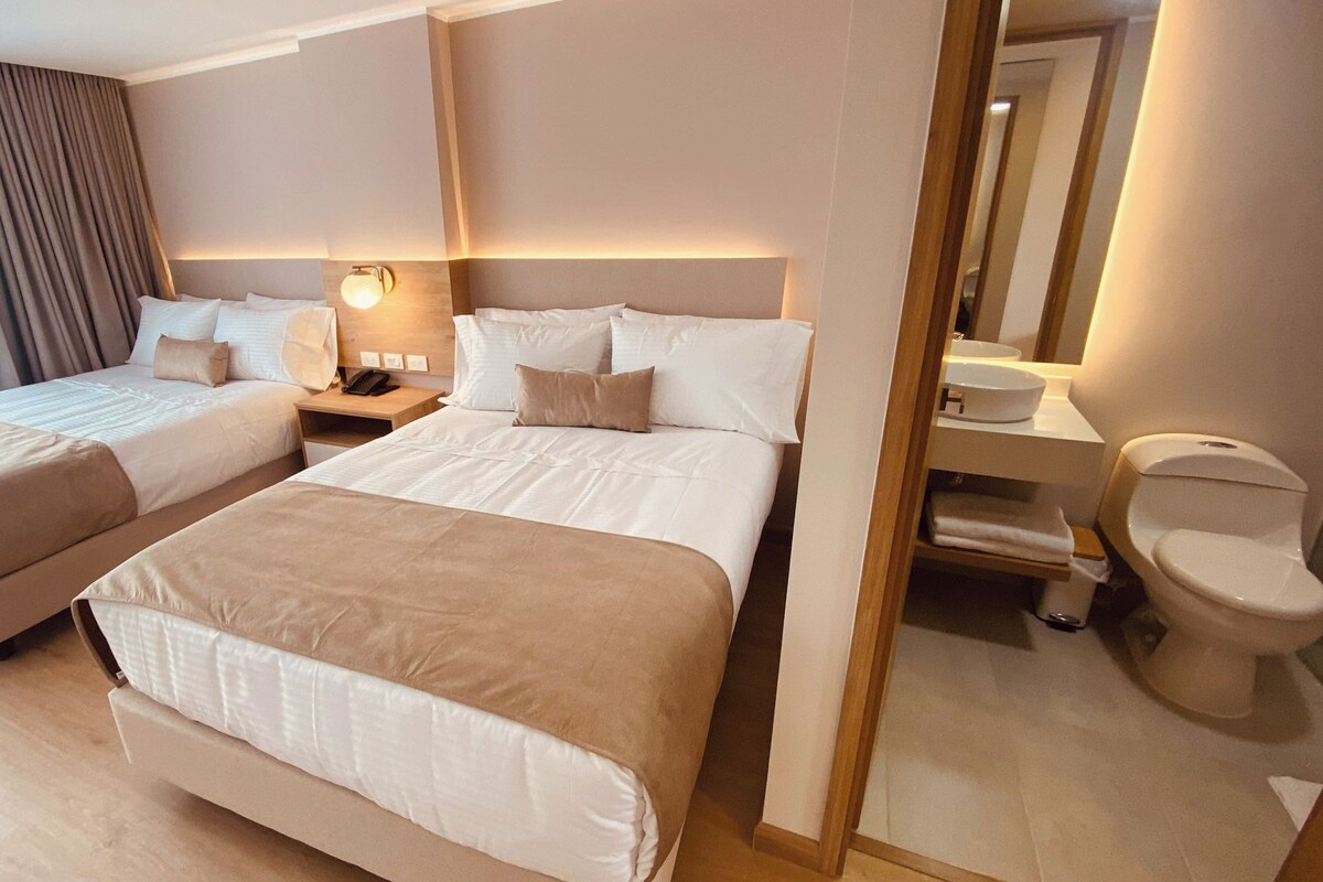 A hotel room with two beds, a bedside table with a lamp, a mirror, and a toilet.