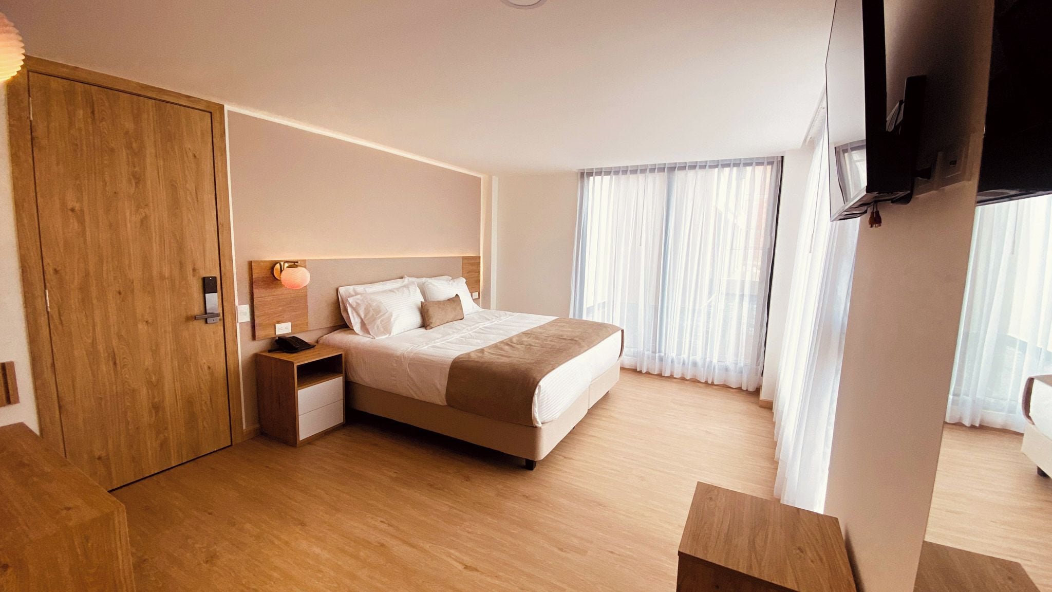 Habitación de hotel con cama, mesita de noche, armario de madera, televisor montado en la pared y cortinas de gasa.