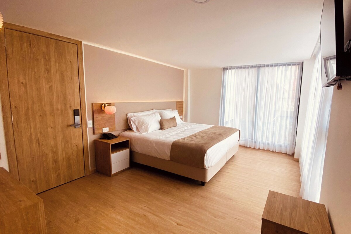 A hotel room with a bed, bedside table, a wooden wardrobe, a TV mounted on the wall, and sheer curtains.