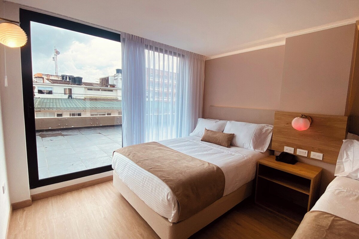 A hotel room with a large window, a bed, a bedside table, and a nightstand.