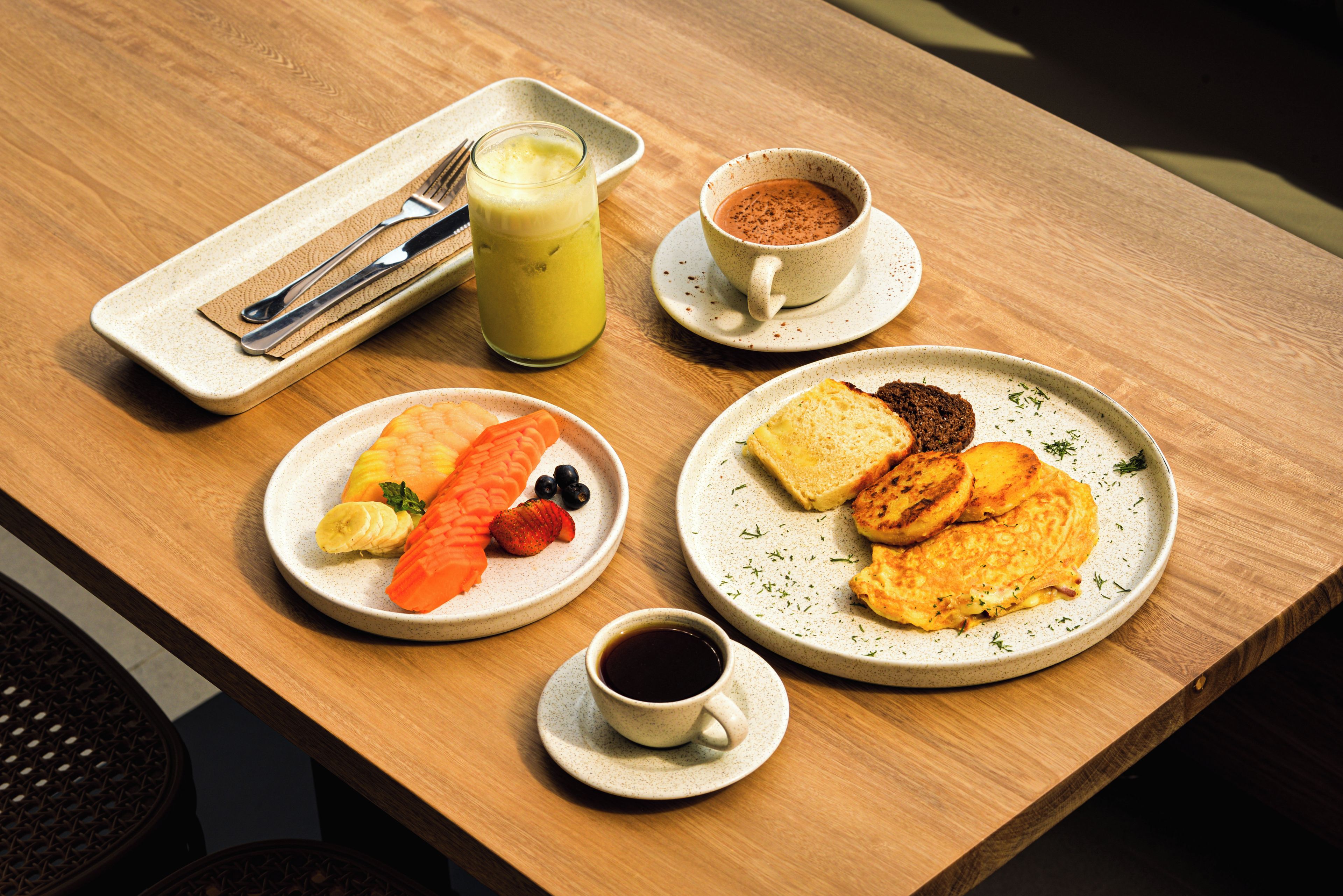 Un desayuno con tostadas, panqueques y fruta, una taza de café, un vaso de jugo y una taza de chocolate caliente sobre una mesa de madera.