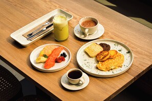Un desayuno con tostadas, panqueques y fruta, una taza de café, un vaso de jugo y una taza de chocolate caliente sobre una mesa de madera.