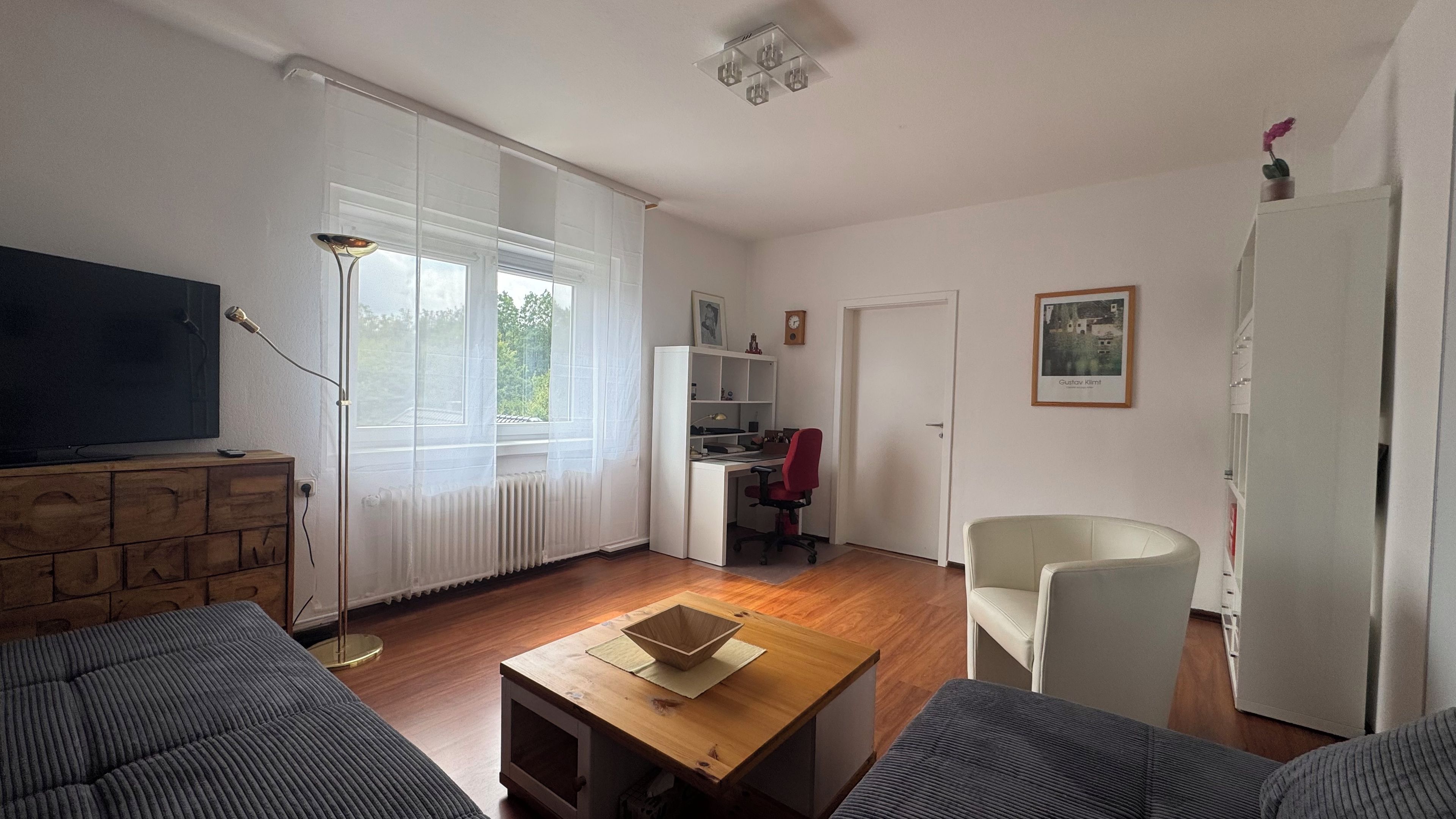 Ein Wohnzimmer mit Fernseher, einem Holztisch, einem roten Bürostuhl und einem weißen Schreibtisch mit Computer.