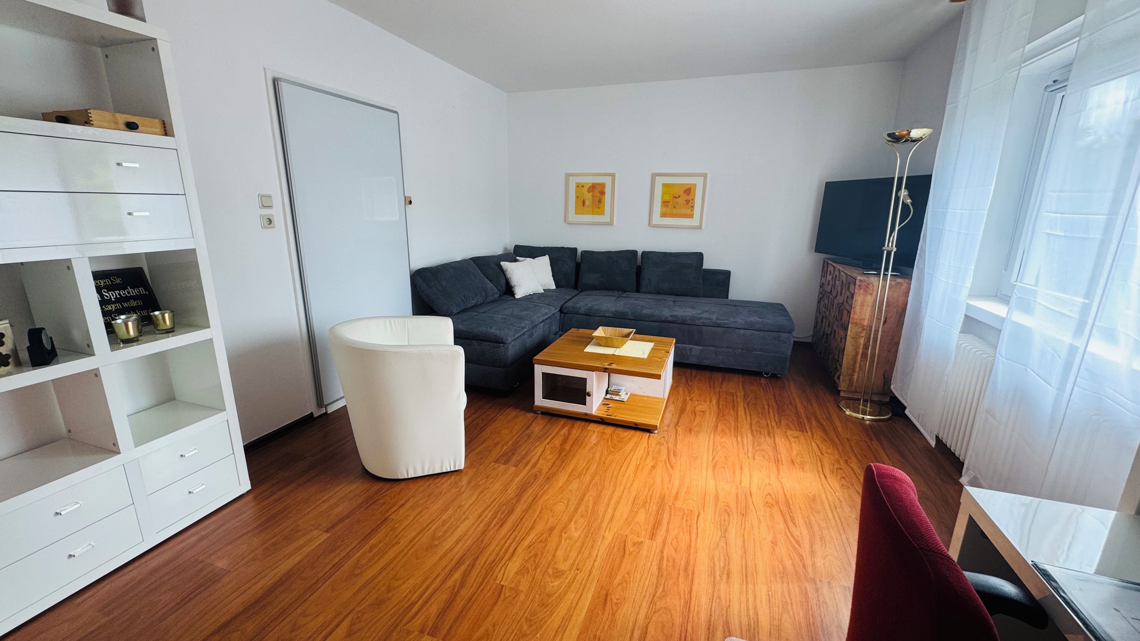 Ein modernes Wohnzimmer mit einer Eckcouch, einem weißen Hocker, einem Couchtisch aus Holz und einem Bücherregal.