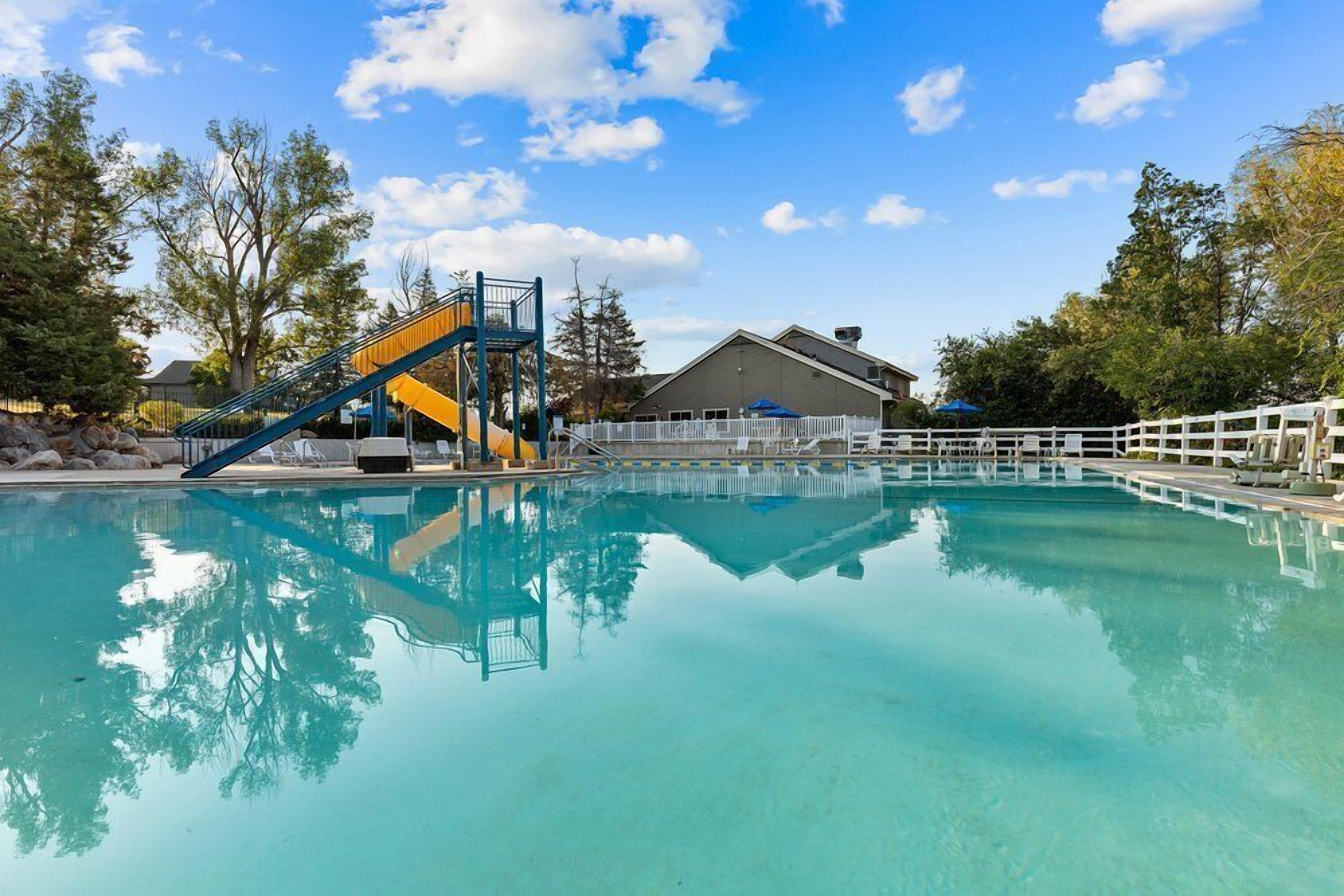 Une piscine aux eaux cristallines, avec un toboggan bleu et jaune, entourée d’arbres et d’un bâtiment délimité par une clôture blanche.