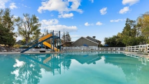 A clear swimming pool with a blue and yellow slide, surrounded by trees and a building with a white fence.