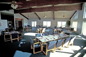 Uma sala de jantar ampla, com várias mesas e cadeiras, tetos altos com vigas aparentes e grandes janelas que permitem entrada de luz natural.