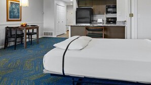 A hotel room with a bed, a desk, a chair, a kitchenette, and a painting on the wall.