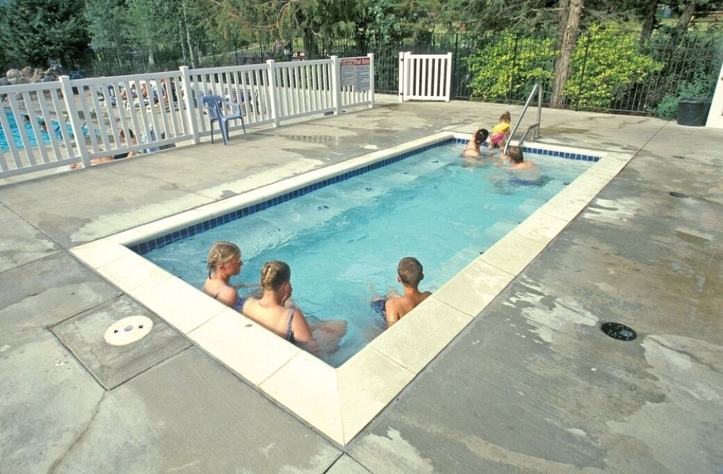 A rectangular swimming pool with a white fence, a few people in the water, and a tree-lined background.