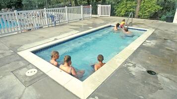 A rectangular swimming pool with a white fence, a few people in the water, and a tree-lined background.