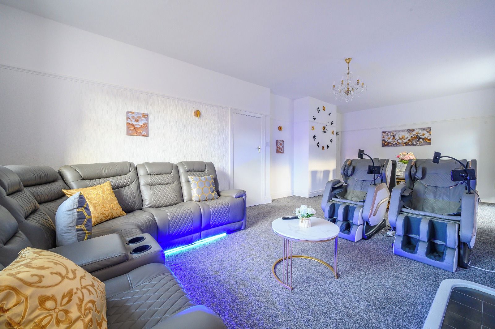 Una sala de estar moderna con un sofá gris, sillones de masaje, una mesita redonda y un reloj de pared.