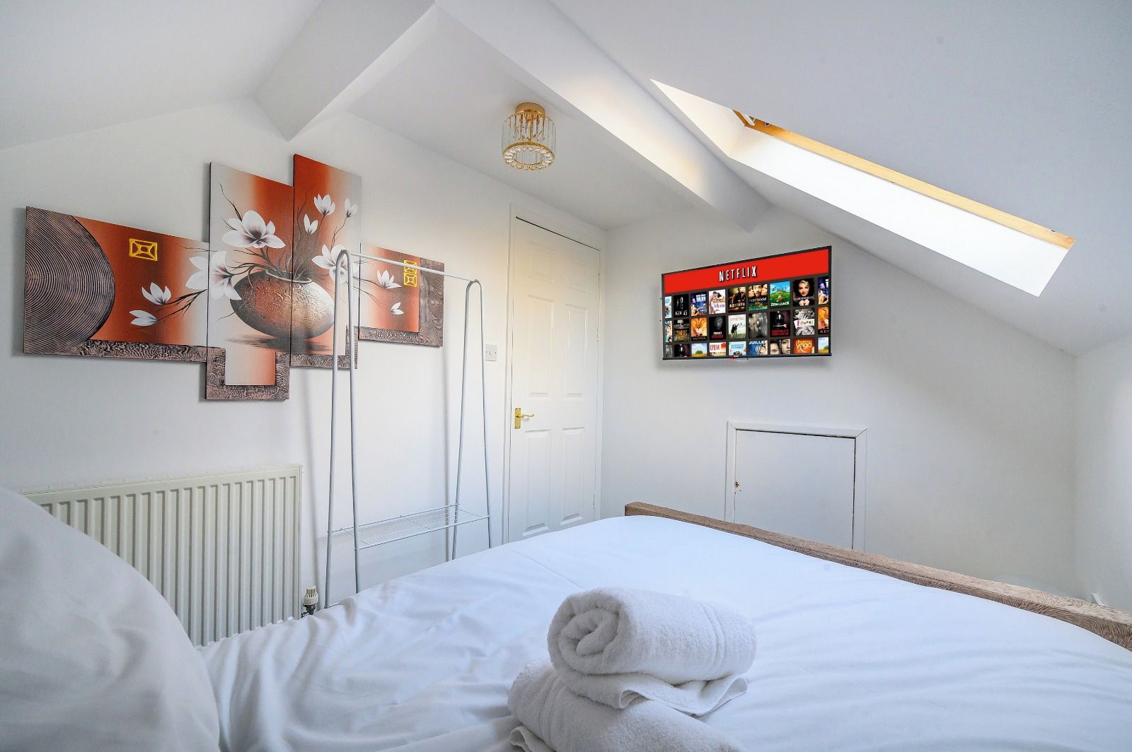 A bedroom with a skylight, a bed with white linens, a wall with a Netflix sign, and artwork.