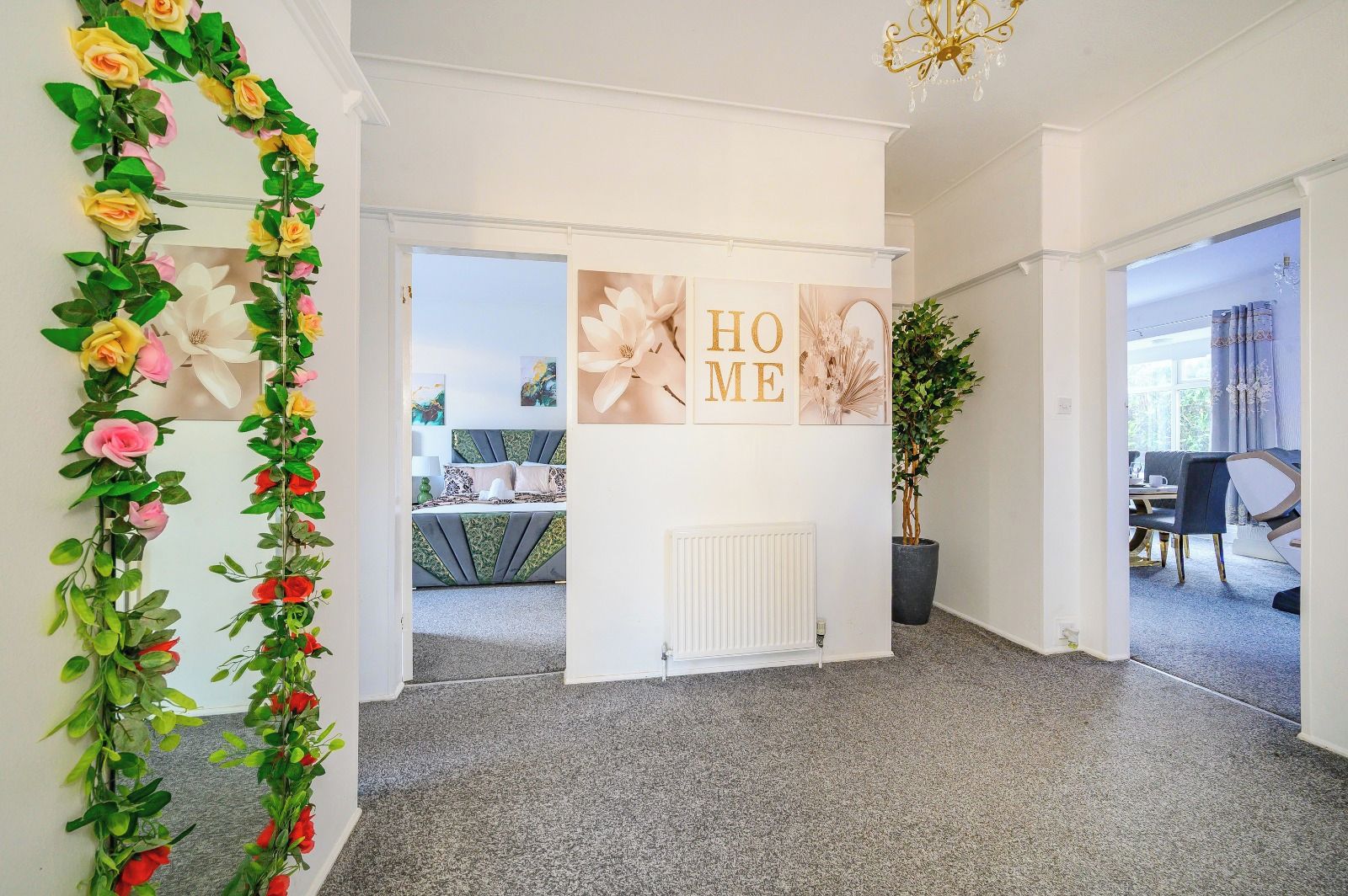 Een gang met een decoratieve bloemenstelling, een witte muur met een 'HOME' bord, en een uitzicht op een eetgedeelte.