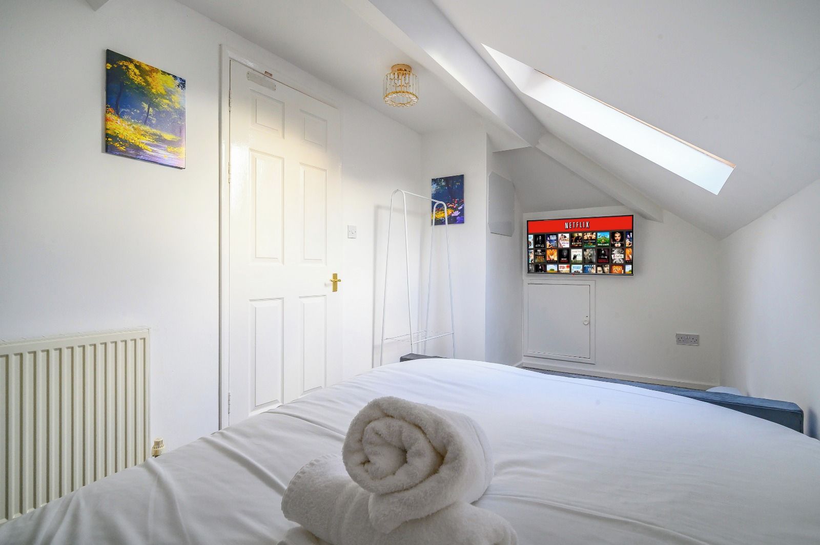 A bedroom with a bed, a radiator, a door, a wall-mounted TV, and a skylight.