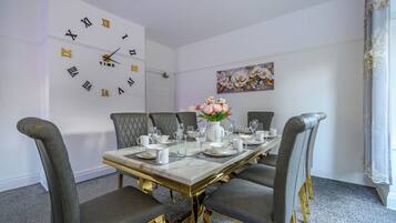 A dining room with a marble table, grey upholstered chairs, a wall clock, and a floral painting.