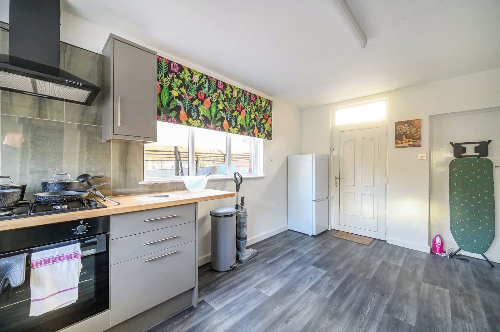 A modern kitchen with a gas stove, oven, and a stainless steel range hood. There is a window with a view of the outside, a white refrigerator, and a green ironing board.