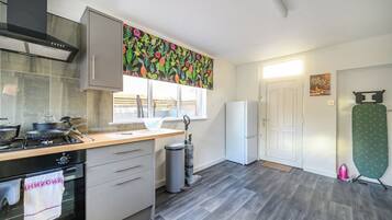 A modern kitchen with a gas stove, oven, and a stainless steel range hood. There is a window with a view of the outside, a white refrigerator, and a green ironing board.