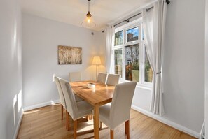 A dining room with a wooden table, six chairs, a lamp, and a window with curtains. - Period 2-Bed Home · Sleeps 4 · Parking · Walk to Wimbledon Station (Greater London)