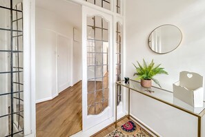 Un couloir avec une porte en verre, un miroir au mur, une petite plante en pot et une table en verre sur laquelle se trouve un objet blanc.