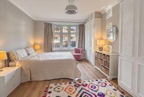 A bedroom with a bed, bedside lamps, a window with curtains, a pink chair, a wooden floor, and a patterned rug. - Period 2-Bed Home · Sleeps 4 · Parking · Walk to Wimbledon Station (Greater London)