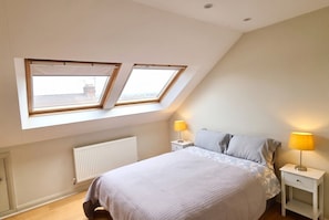 Ein Schlafzimmer mit schrägen Wänden, einem Bett mit gemustertem Überwurf, zwei Nachttischlampen und einer Heizung.