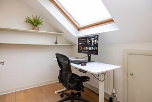 A room with a sloped ceiling, a desk with a computer, a chair, a small plant, and a door. - 3-Bed Flat · Sleeps 6 · Workspace · 10-Min Walk to Wimbledon Station (Greater London)