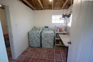 A compact room with a tiled floor, a white door, a window with a wooden beam ceiling, and a shelf with various items. - The Buck Island Overlook | Sleeps 12 (Christiansted)