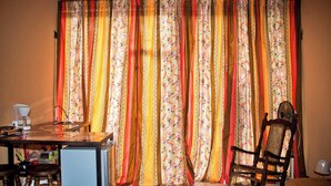Una habitación con comedor, una silla de madera, una mesa con cafetera y una cortina de estampado floral.
