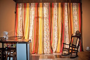 A room with a dining area, a wooden chair, a table with a coffee maker, and a curtain with a floral pattern.