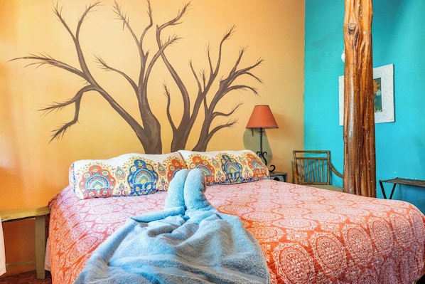 Una cama con ropa de cama estampada, un mural de un árbol en la pared, una lámpara y una silla.