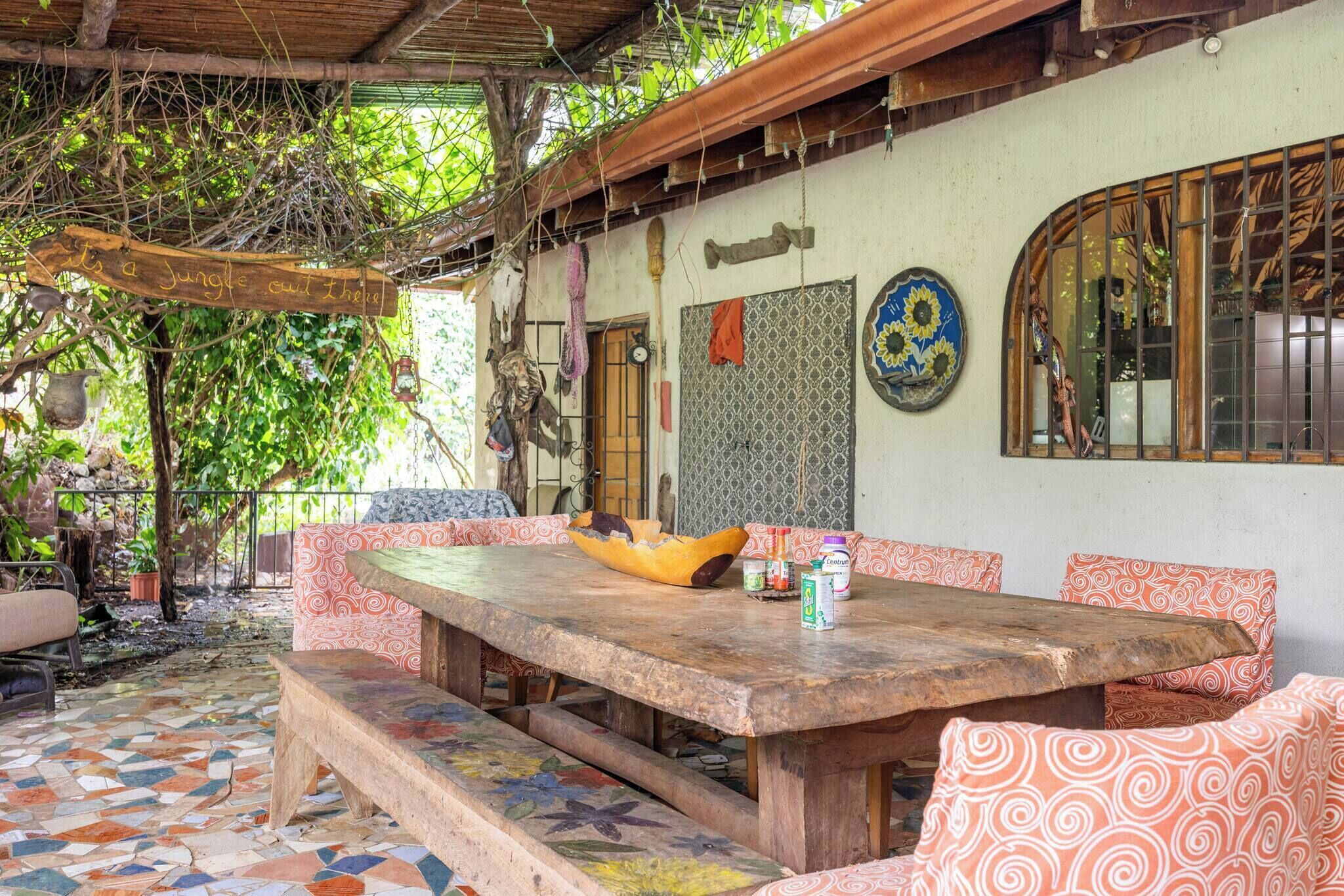 An outdoor dining area with a wooden table and benches, surrounded by a patterned floor and a rustic wall.