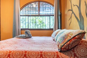 Un lit avec une couvre-lit à motifs et deux oreillers, une fenêtre donnant sur un paysage verdoyant et un mur orné d’une fresque représentant un arbre.