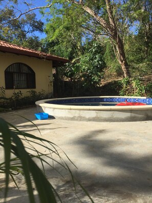 Une piscine avec un bord carrelé, un récipient en plastique bleu et une maison avec un toit rouge et une fenêtre en arc de cercle.