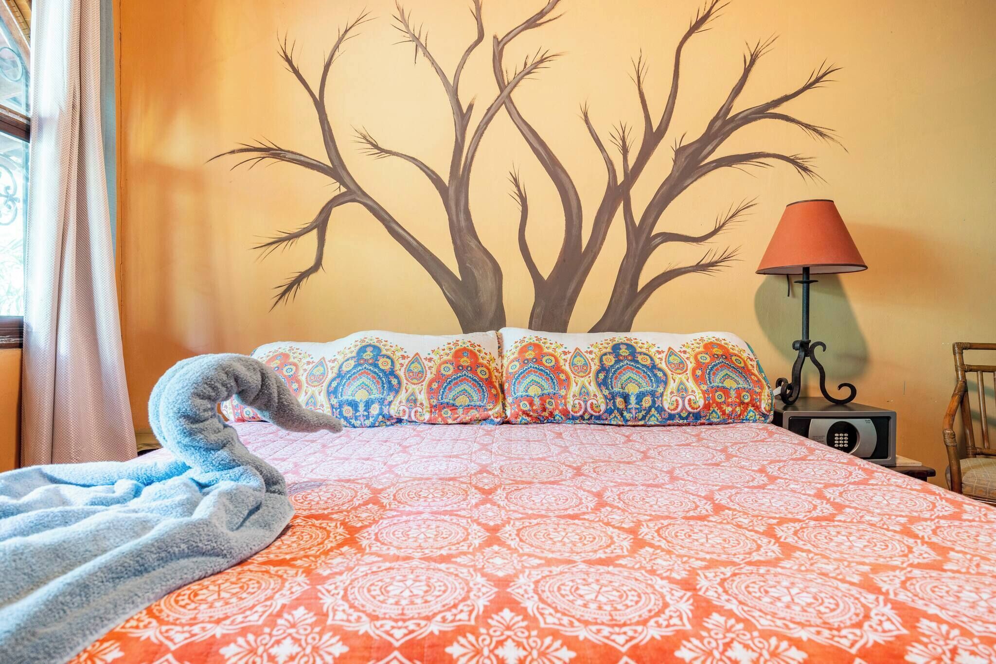 Ein Bett mit gemusterten Betttextilien, eine Wandmalerei eines Baumes, eine Lampe und ein Sessel.