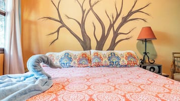Ein Bett mit gemusterten Betttextilien, eine Wandmalerei eines Baumes, eine Lampe und ein Sessel.