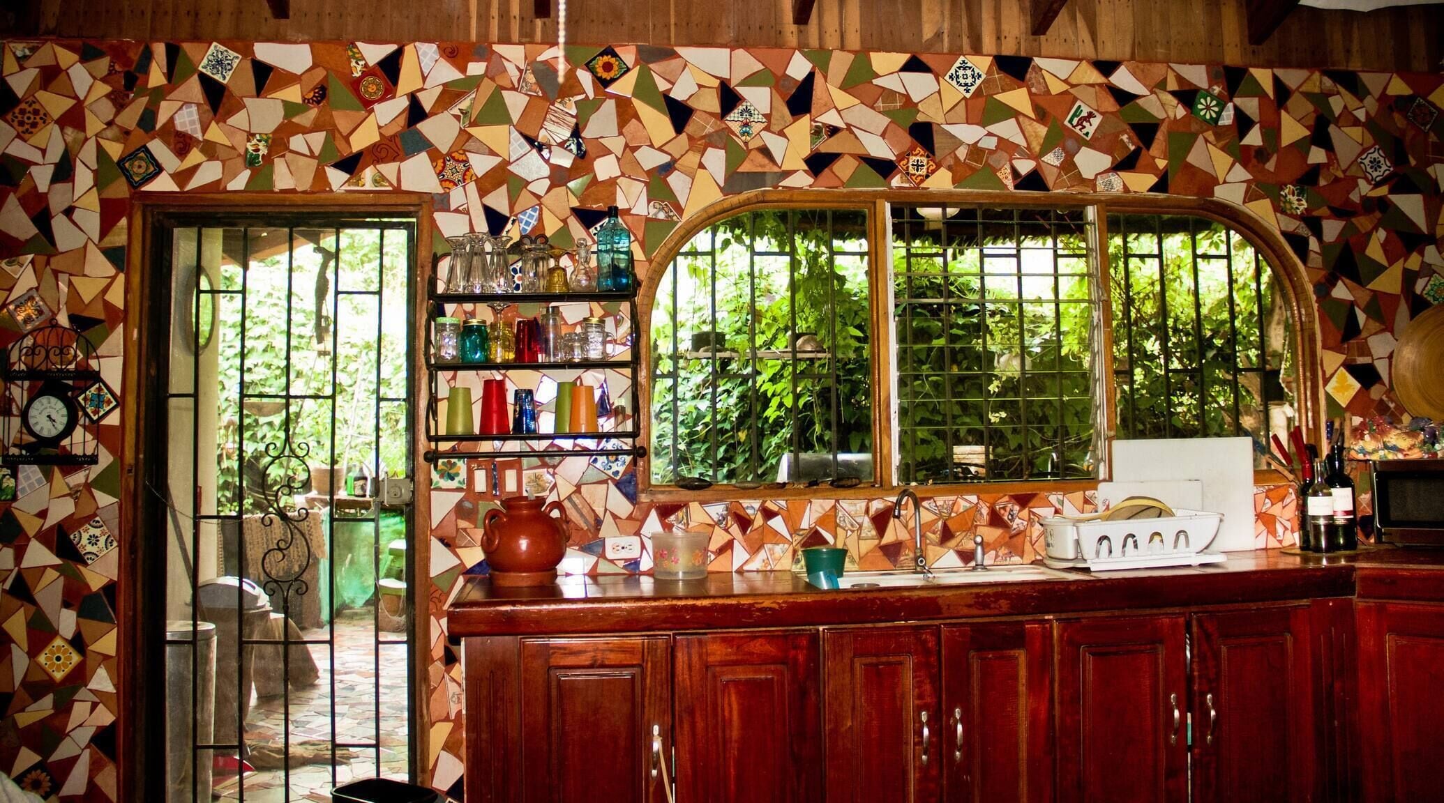 A kitchen with a mosaic backsplash, wooden cabinets, a window with a view of greenery, and a shelf with various kitchen items.