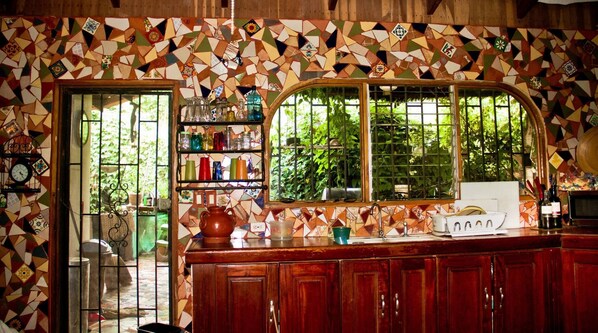 Uma cozinha com painel de backsplash em mosaico, armários de madeira, uma janela com vista para vegetação e uma prateleira com diversos utensílios de cozinha.