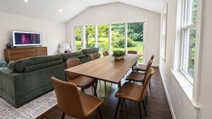 En moderne stue med sofa, spisebord med stole, fjernsyn og et stort vindue med udsigt til grønne omgivelser.