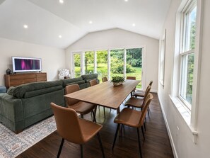 Ein modernes Wohnzimmer mit einer Couch, einem Esstisch mit Stühlen, einem Fernseher und einem großen Fenster mit Blick auf Grünflächen.