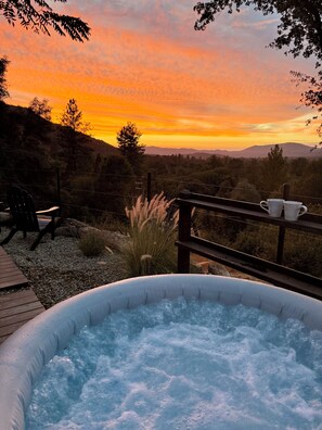 A hot tub with a wooden deck and chairs, overlooking a mountainous landscape during sunset.